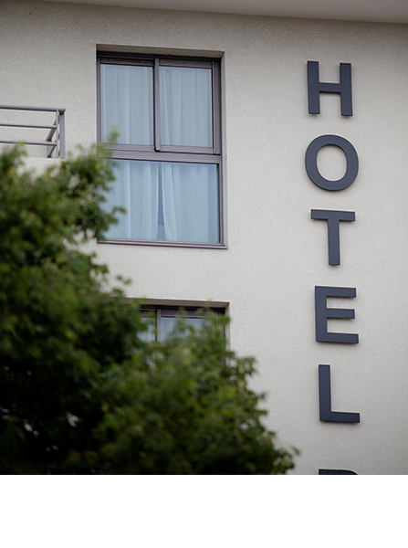 Panneau d'hôtel sur un bâtiment crème avec des fenêtres, partiellement masqué par un arbre vert.