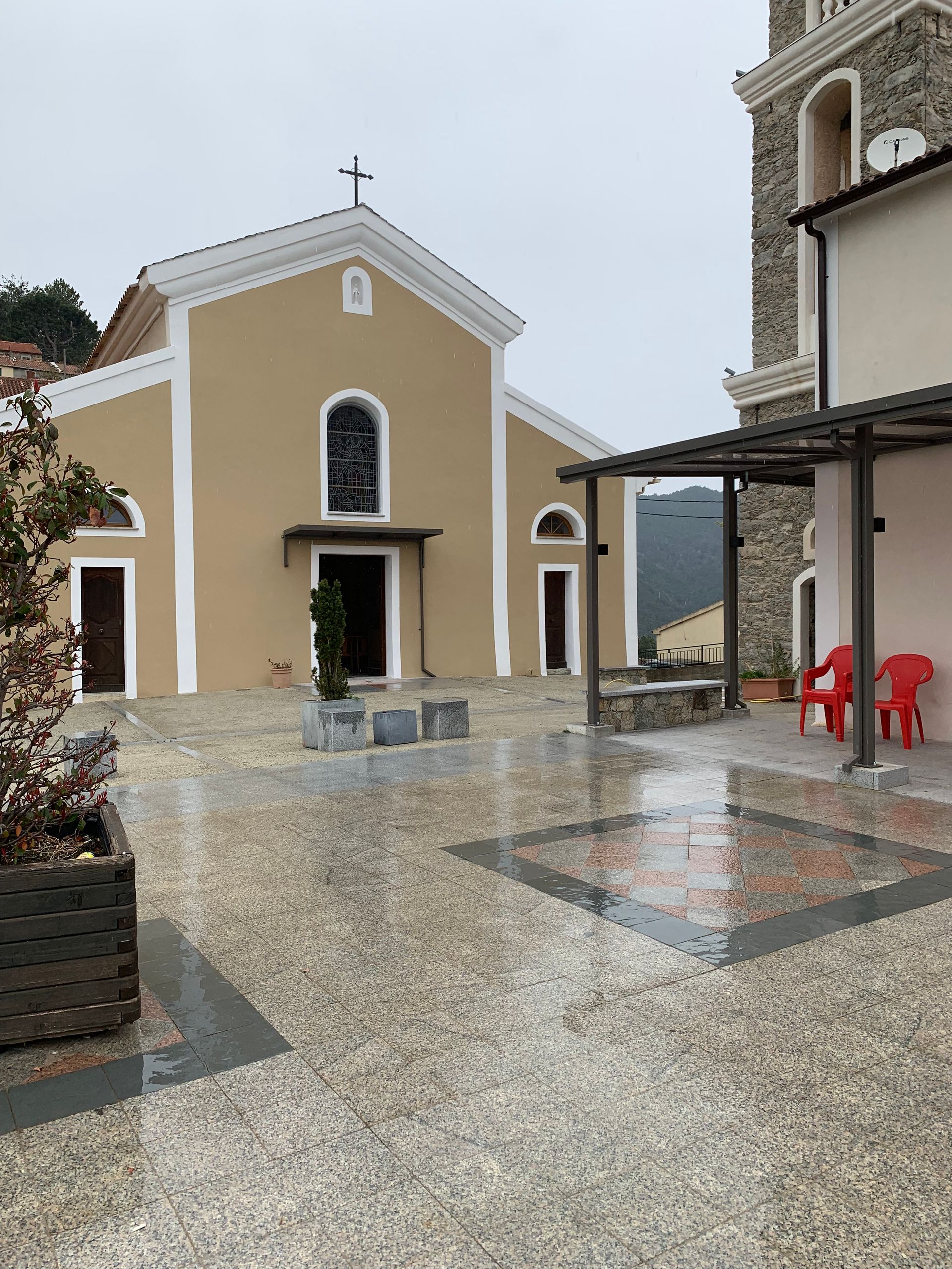 Église à façade beige et croix au sommet, petite cour, pierre grise et clocher à droite.
