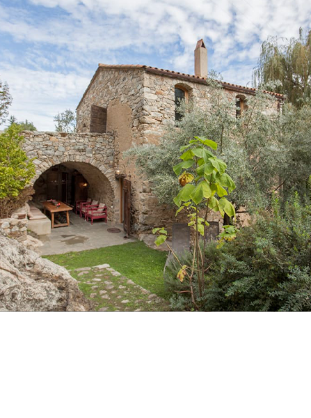 Maison en pierre avec entrée voûtée et patio, entourée de verdure luxuriante.