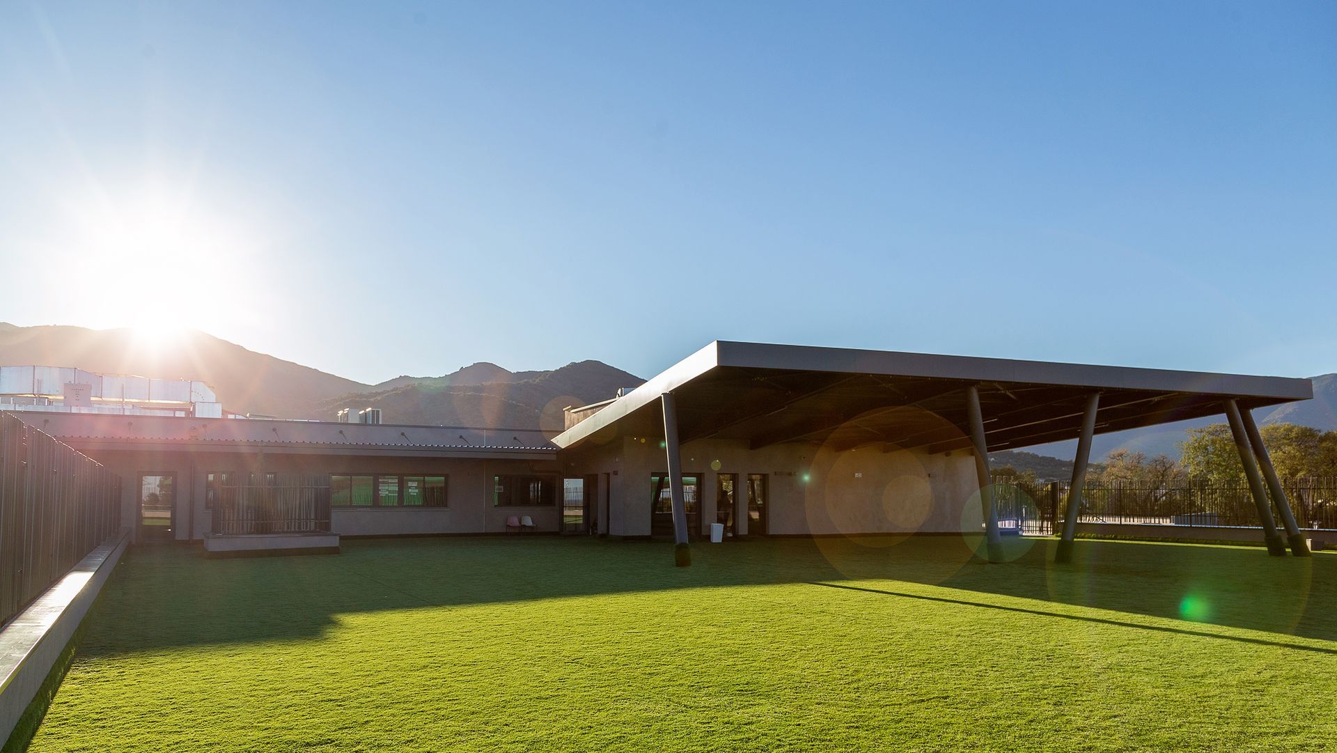Journée ensoleillée avec un bâtiment moderne et bas, une pelouse verte et des montagnes en arrière-plan.