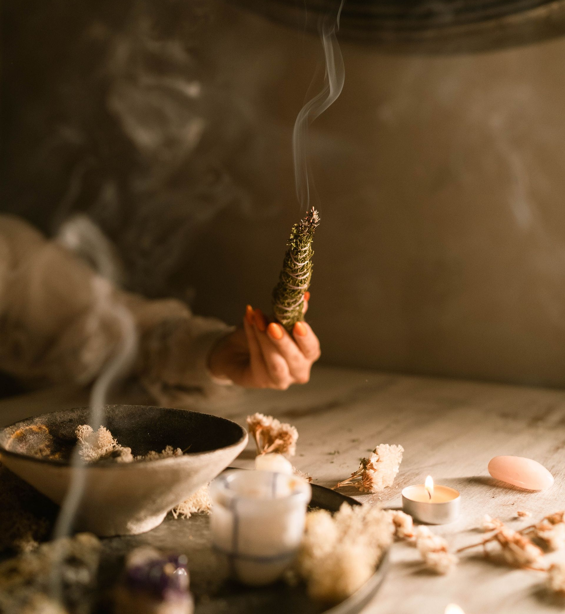 Una mano sostiene un manojo de salvia humeante sobre un altar tenuemente iluminado con velas y cristales.