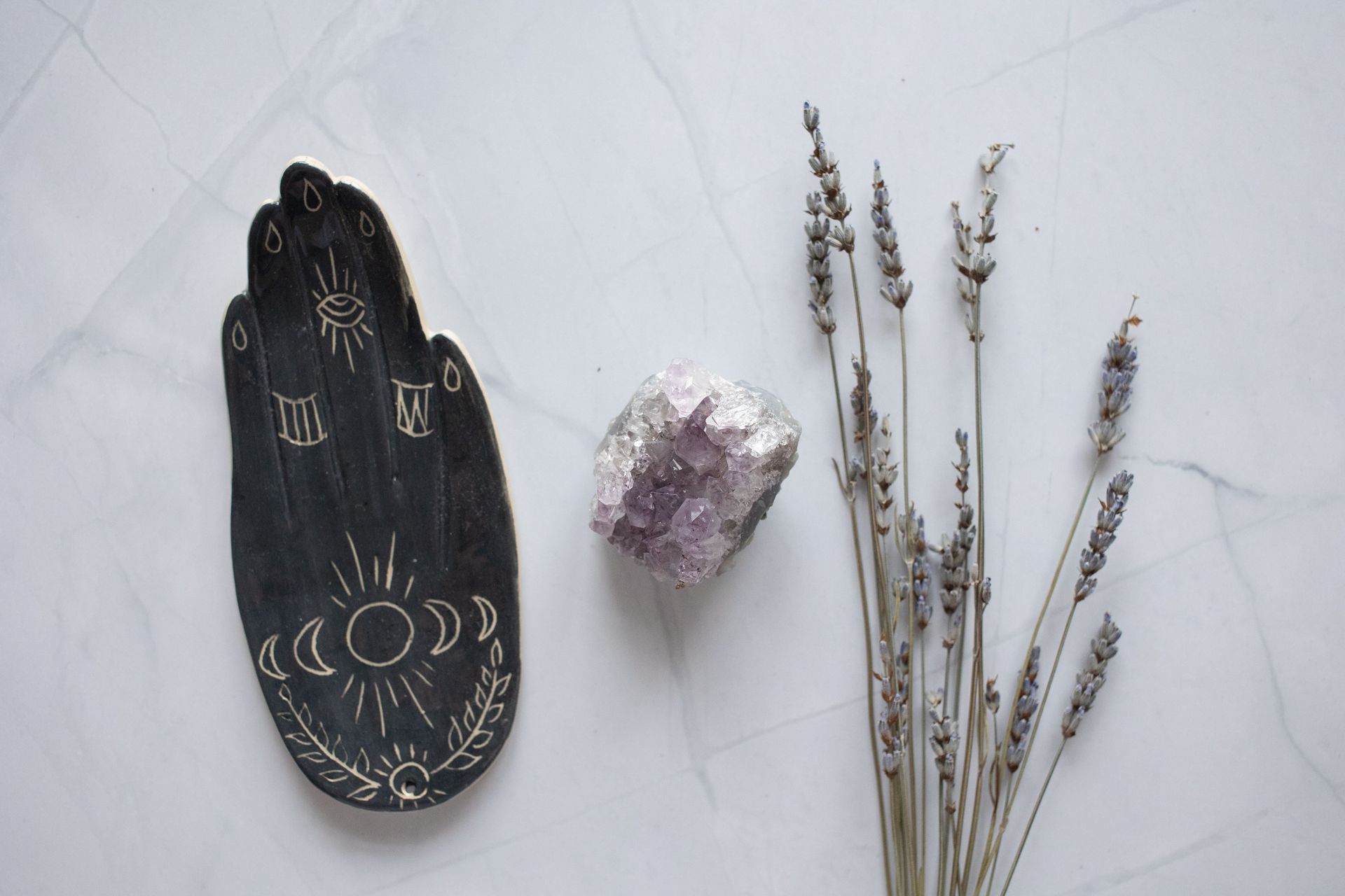 Plato negro con forma de mano, con diseños dorados, cristal morado y lavanda seca sobre una superficie blanca.