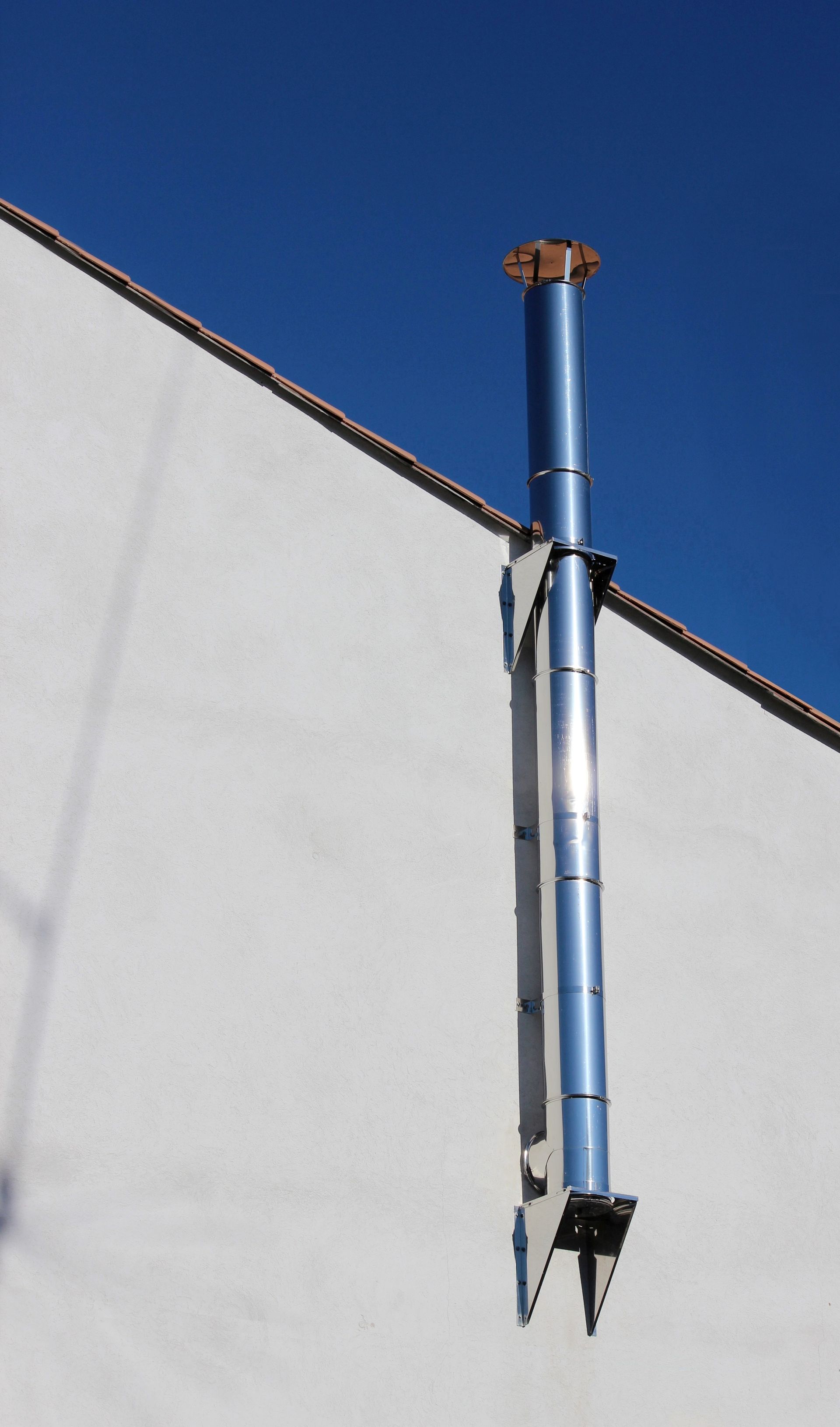 Conduit de cheminée vertical en métal fixé sur un mur blanc sous un ciel bleu.