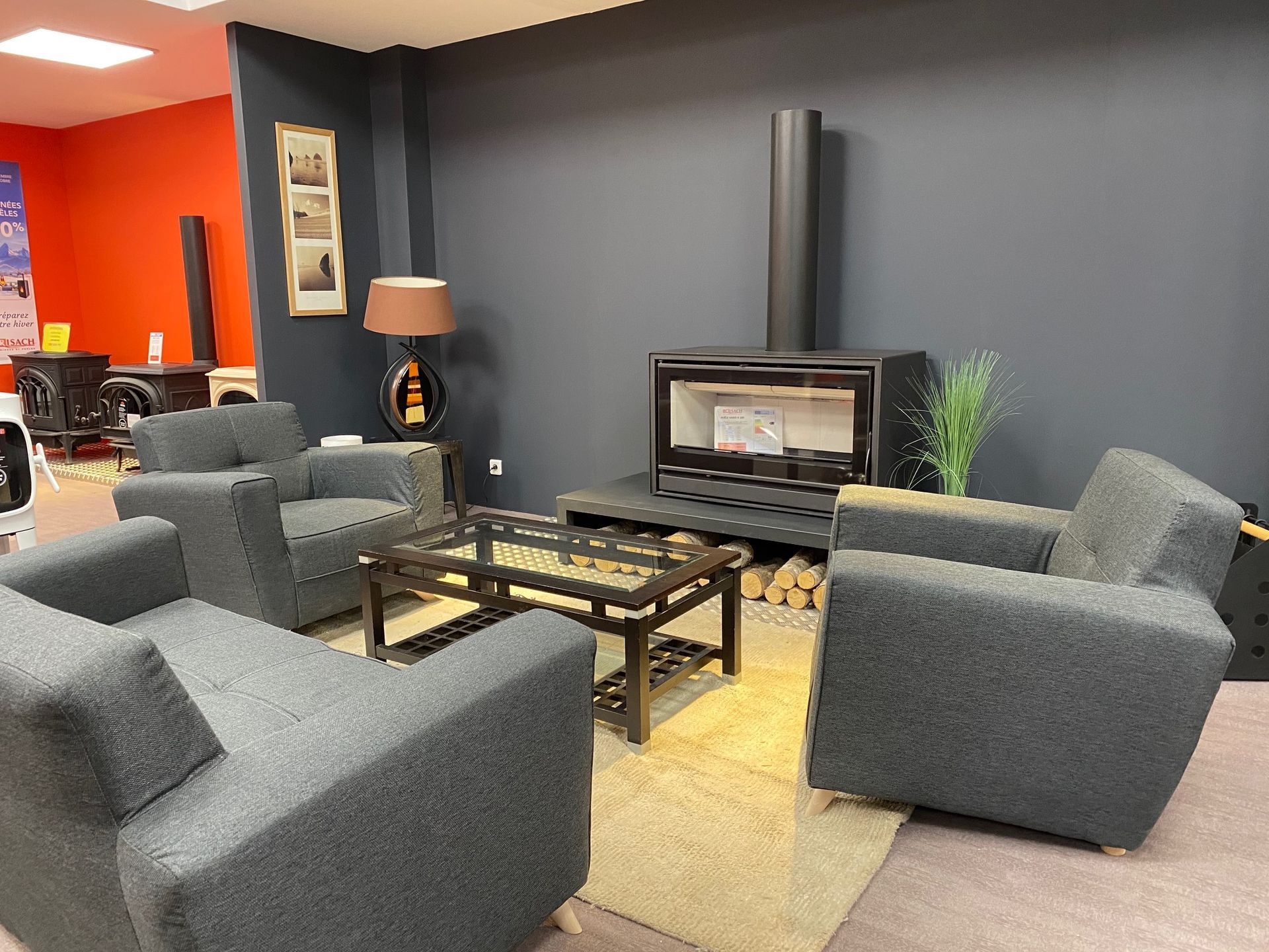 Showroom avec mise en situation avec des fauteuils gris, une table basse en verre et un poêle à bois avec conduit apparent.