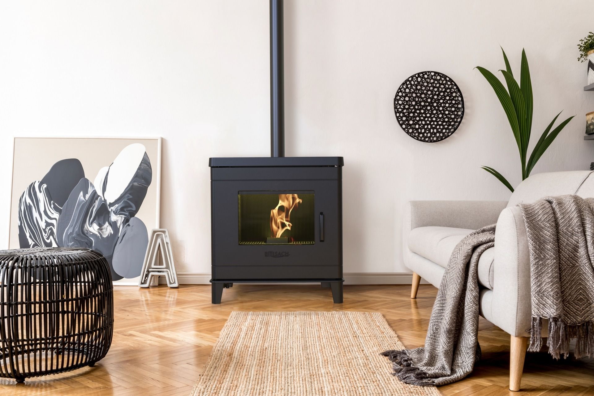 Salon avec un poêle noir, un canapé beige, un fauteuil en osier, une plante et des œuvres encadrées sur un parquet clair.