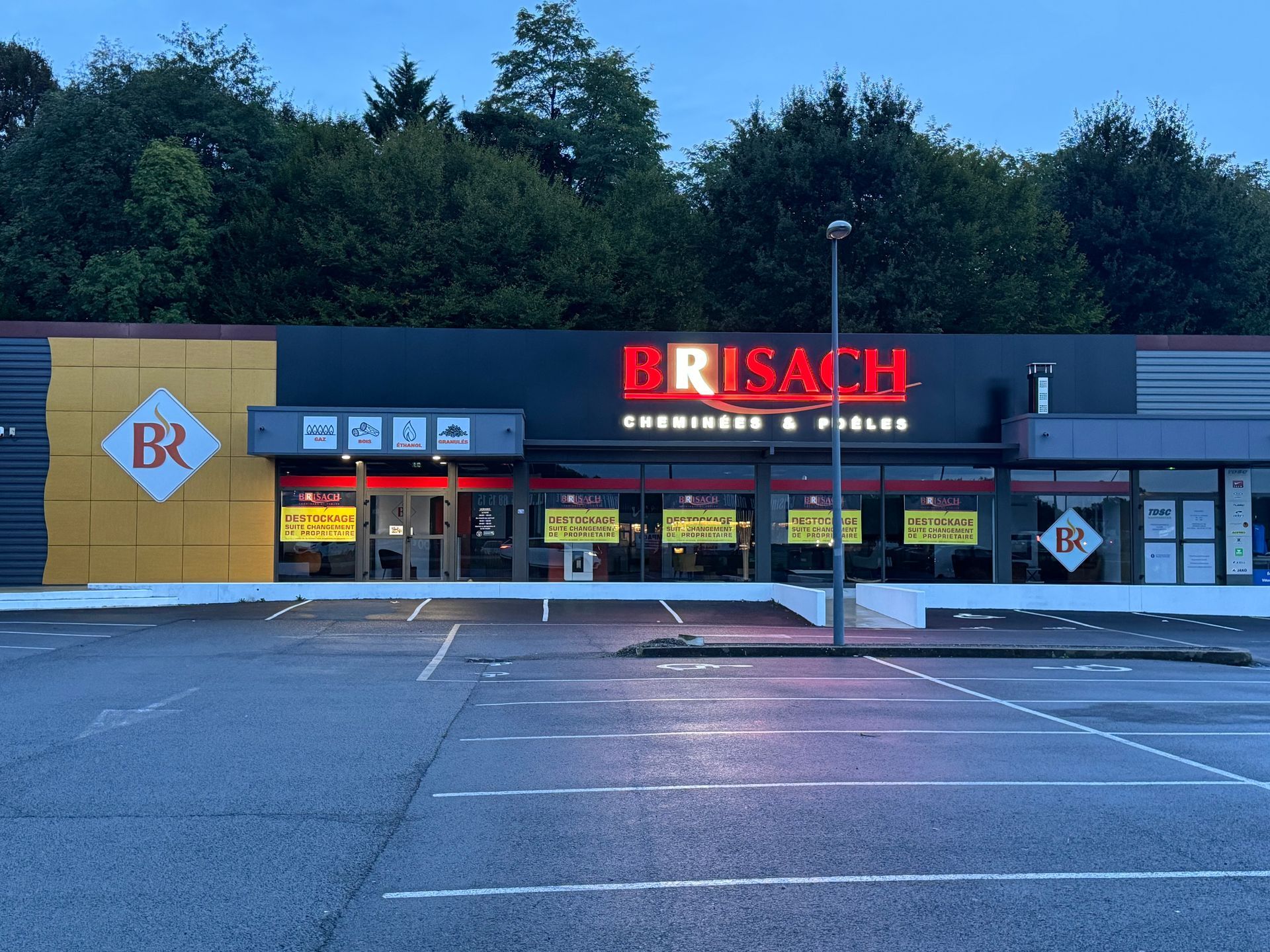 Extérieur du magasin Brisach à Malemort-sur-Corrèze à la tombée de la nuit, avec son enseigne rouge illuminée et un parking vide devant.