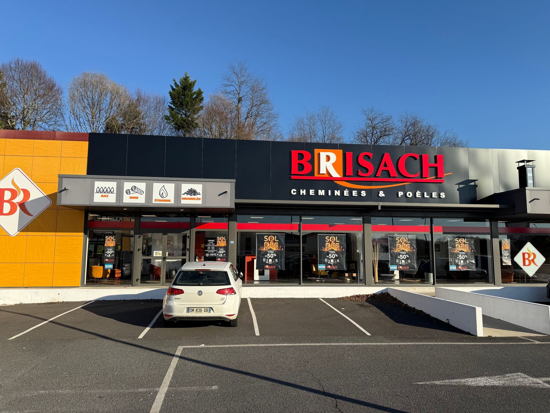 Façade du magasin Brisach avec une enseigne rouge et noire, un parking devant avec une voiture blanche garée.