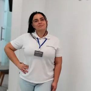 Una mujer con una camisa polo blanca y pantalones azules está parada frente a una pared blanca.
