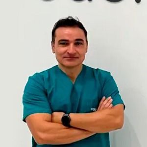 Un hombre con un matorral está de pie con los brazos cruzados frente a una pared blanca.