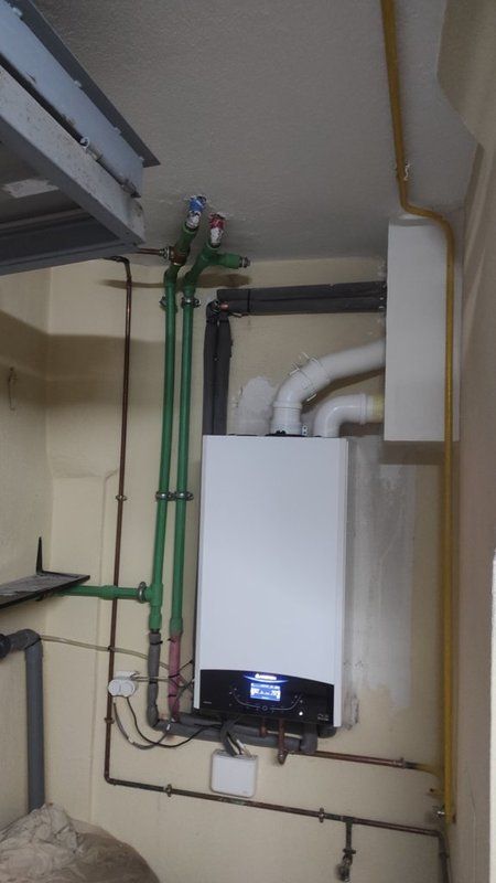 Una caldera blanca está colgada en una pared al lado de tuberías verdes.