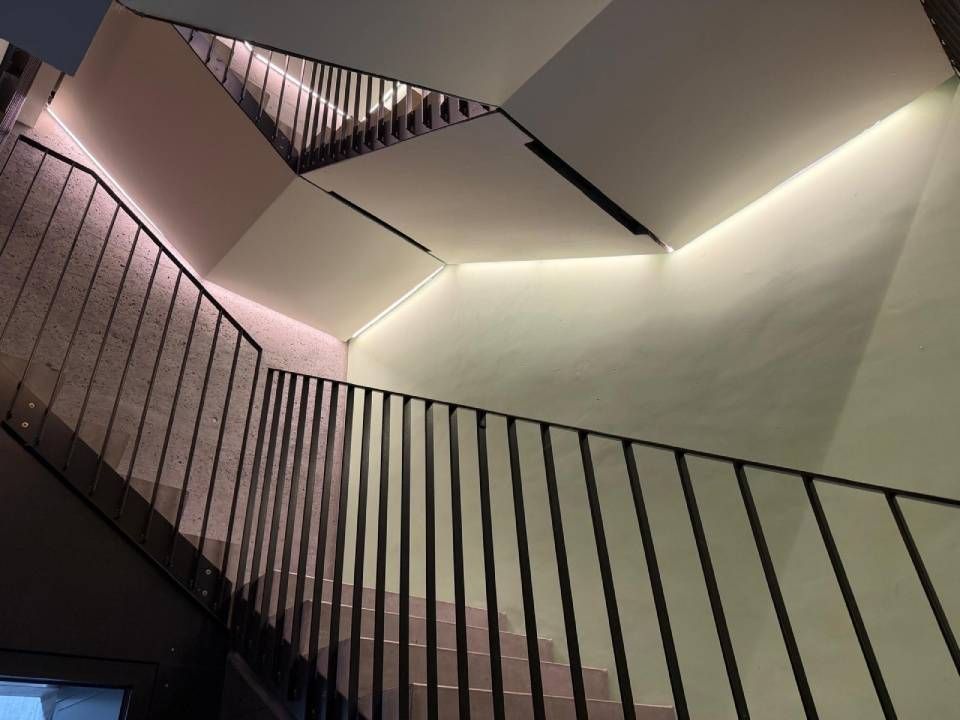 Modernes Treppenhaus mit schwarzem Geländer und schrägen weissen Wänden, beleuchtet von sanftem Deckenlicht.