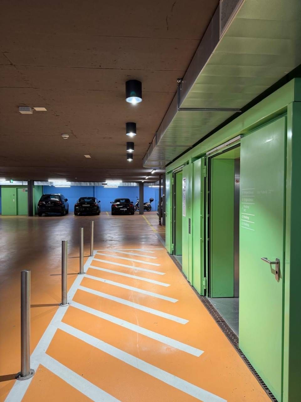 Überdachtes Parkhaus mit grünen Türen und orange markiertem Gehweg.