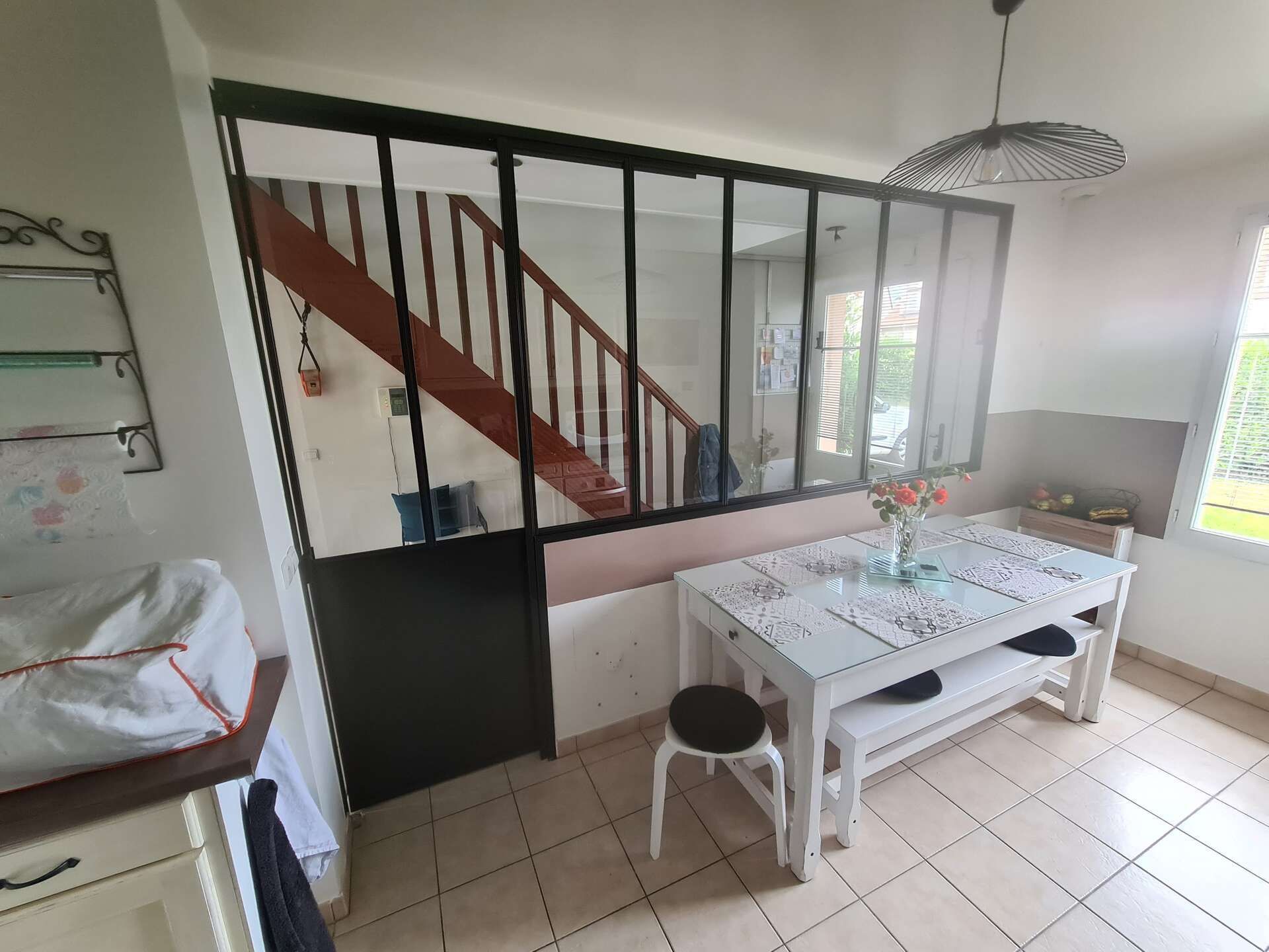 Salle à manger lumineuse avec table blanche, tabourets, sol carrelé et séparation d'escalier par une verrière.