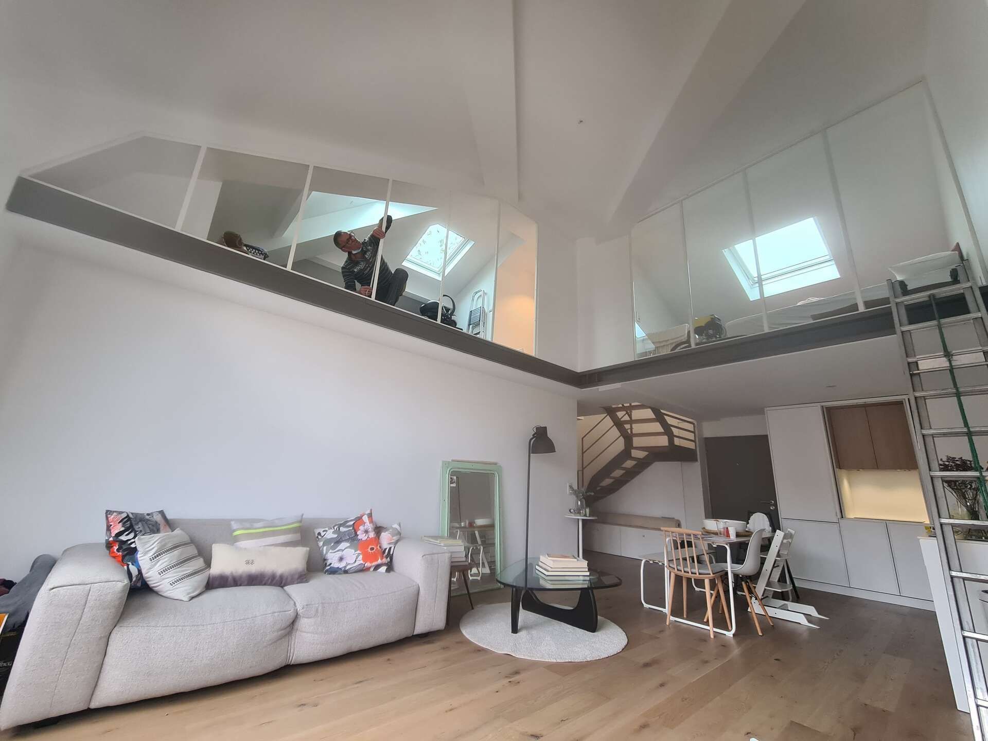 Salon en mezzanine avec canapé gris, escalier, table à manger et puits de lumière.