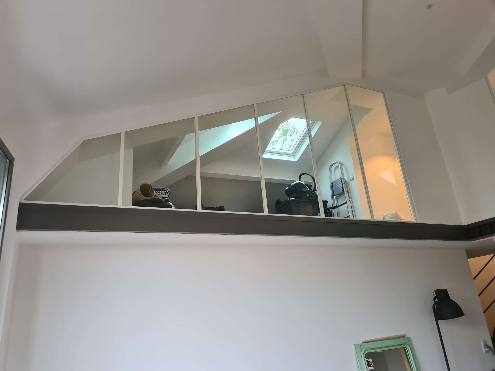 Chambre en mezzanine avec puits de lumière, plafond mansardé et lit derrière une cloison vitrée au-dessus d'un mur blanc.