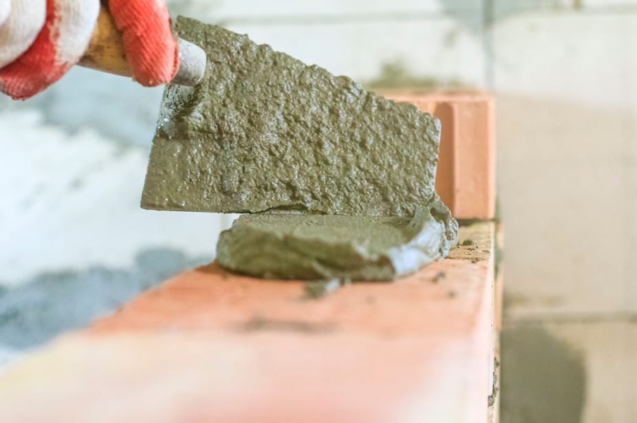 Una mano enguantada utiliza una llana para aplicar mortero húmedo sobre un ladrillo rojo.