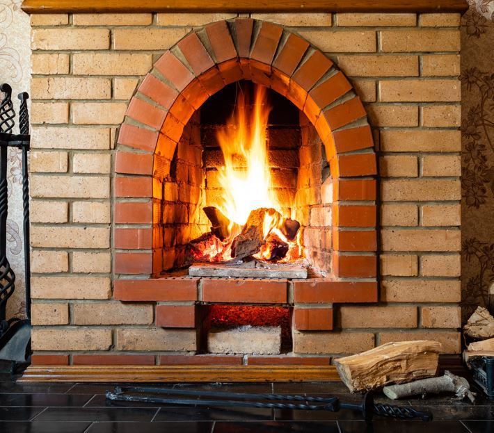 Una chimenea de ladrillo con un fuego brillante ardiendo en su interior, y una pequeña pila de leña en el suelo cerca.
