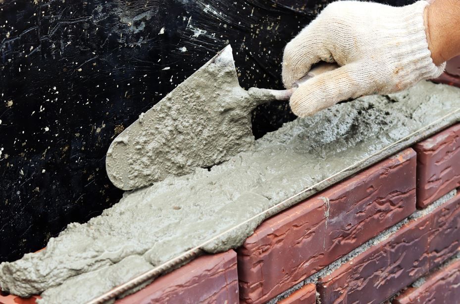 Una mano enguantada utiliza una llana para aplicar mortero húmedo sobre una pared de ladrillos en construcción.
