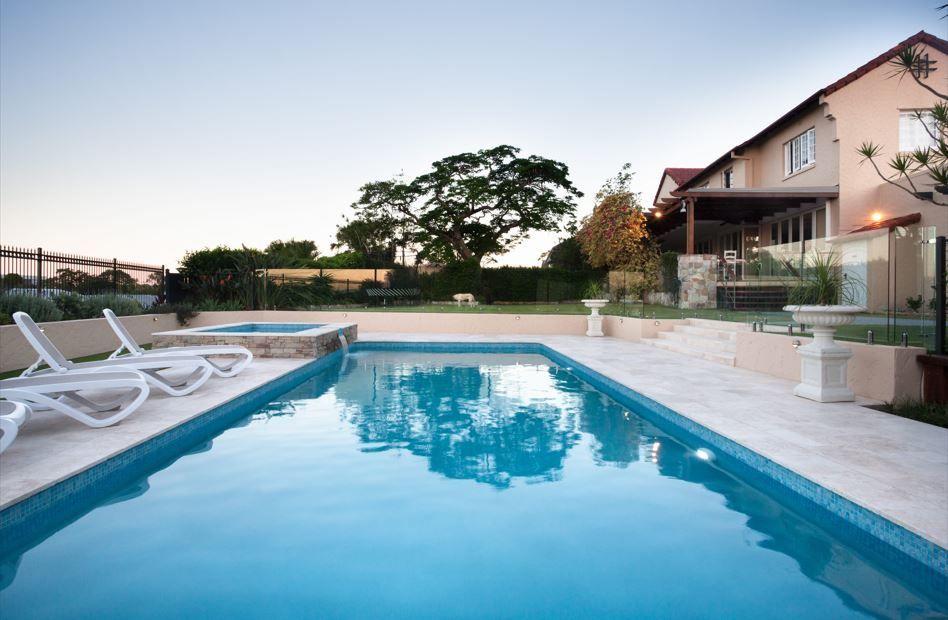 Una piscina rectangular con tumbonas blancas en un patio de piedra junto a una casa al atardecer.
