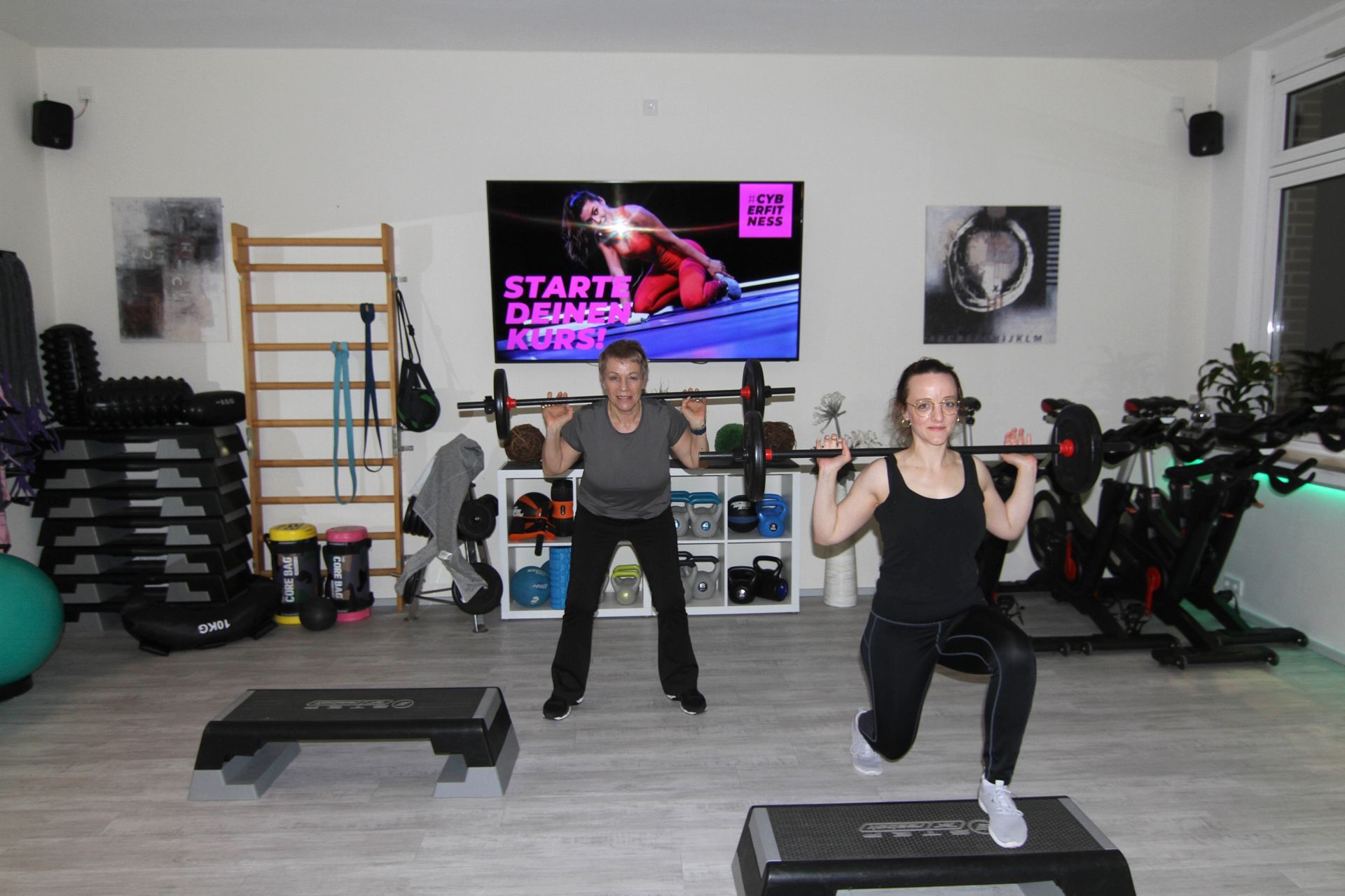 Zwei Frauen machen in einem Fitnessstudio vor einem Fernseher Kniebeugen.