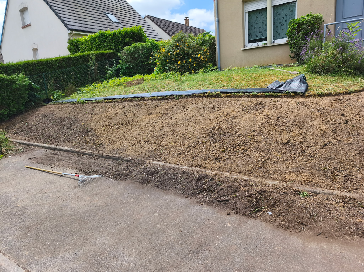 Talus de terre fraîchement creusé le long d'un trottoir et d'une pelouse résidentielle, avec une maison et une haie en arrière-plan.
