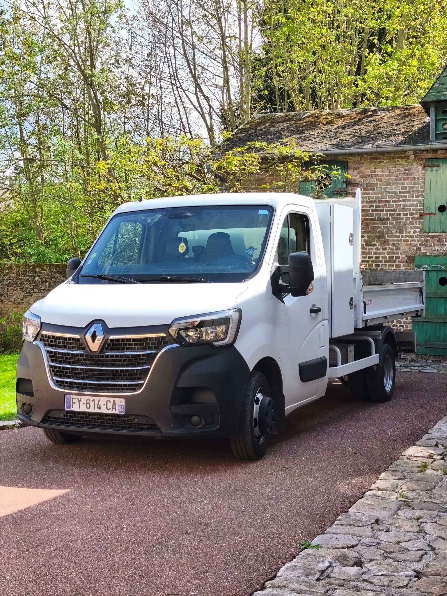 Une camionnette Renault blanche est garée dans une allée, à côté d'un bâtiment en briques et de verdure.