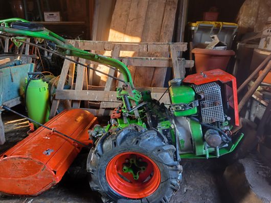 Tracteur vert et orange avec tondeuse à gazon garé dans un hangar en bois encombré