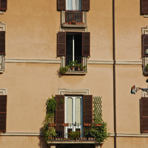 Façade de bâtiment beige avec plusieurs fenêtres et balcons, certains avec des volets bruns ouverts et de la verdure.