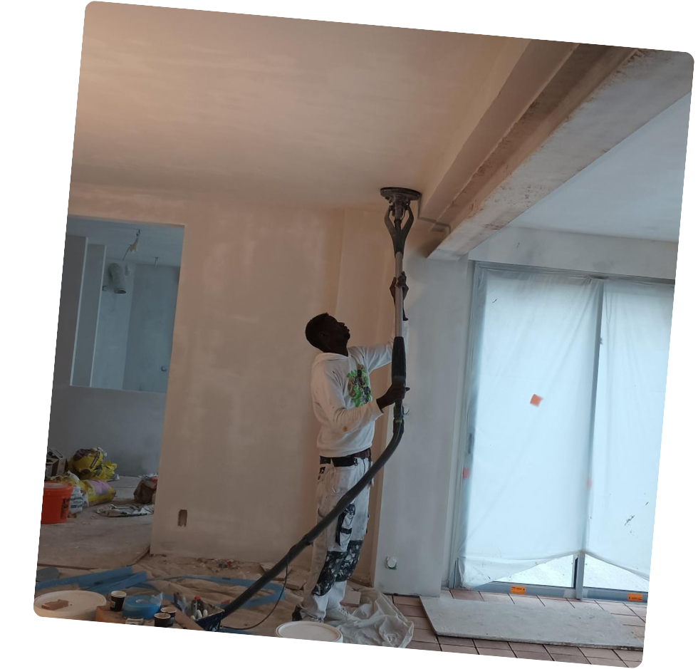 Un homme ponce un plafond blanc avec une ponceuse à long manche. Aménagement intérieur avec protections sur les fenêtres.