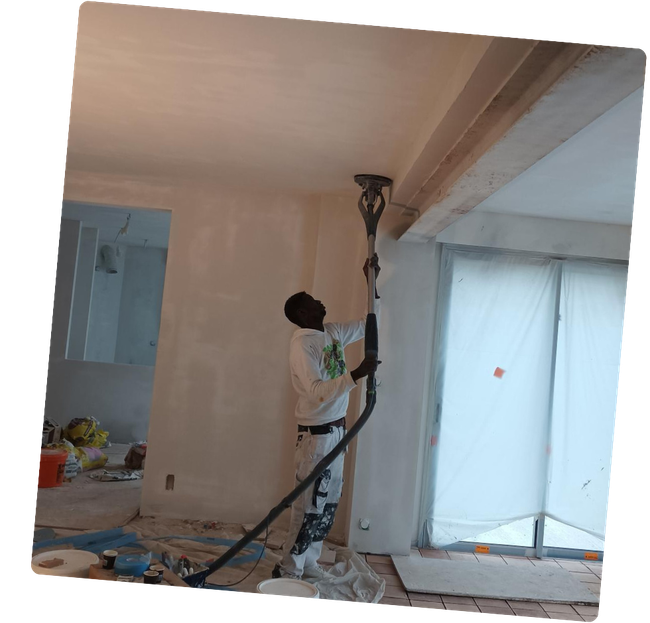 Un homme ponce un plafond blanc avec une ponceuse à long manche. Aménagement intérieur avec protections sur les fenêtres.