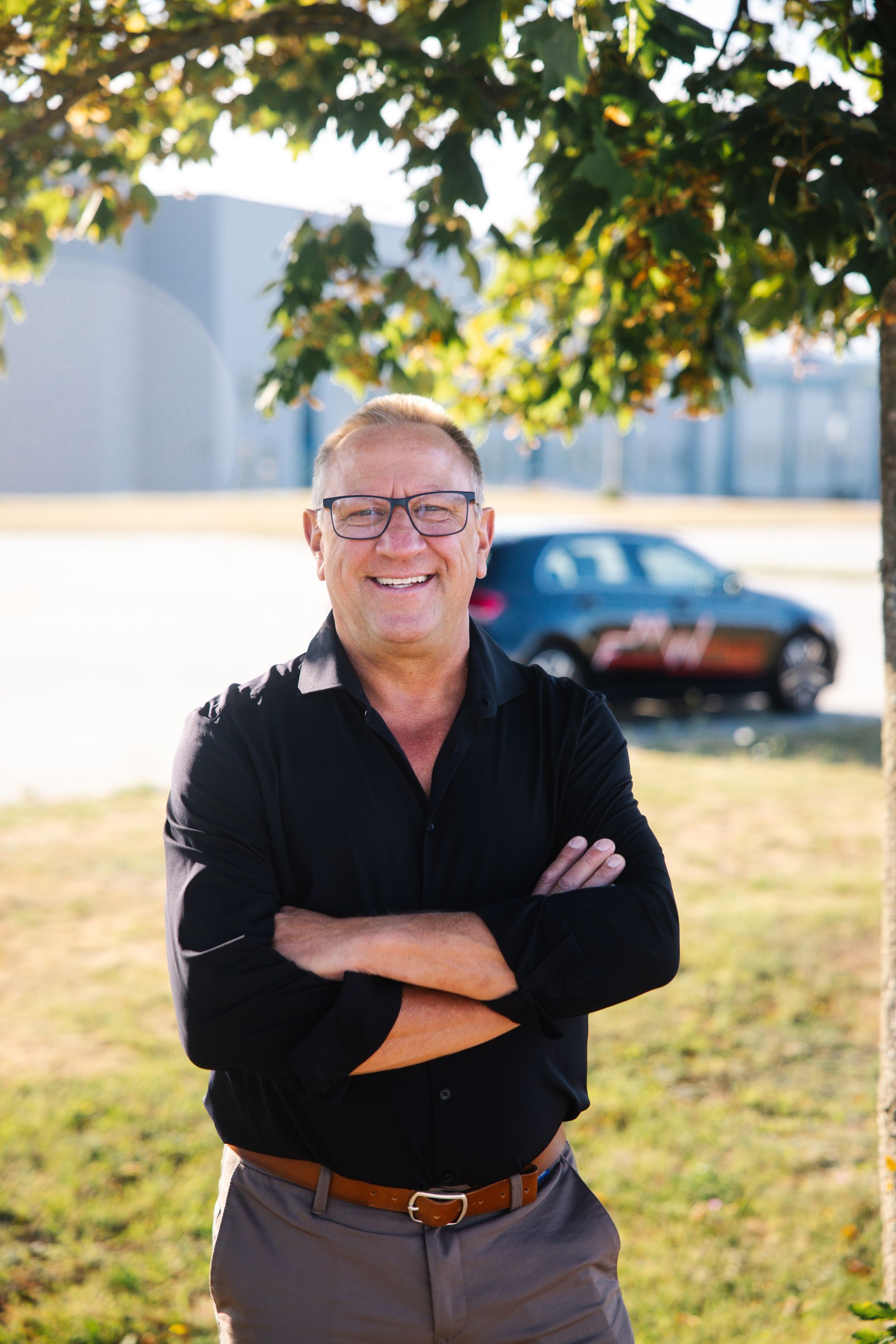 Mann mit Glatze, schwarzem Hemd, verschränkten Armen, lächelt. Auto im Hintergrund. Draußen.