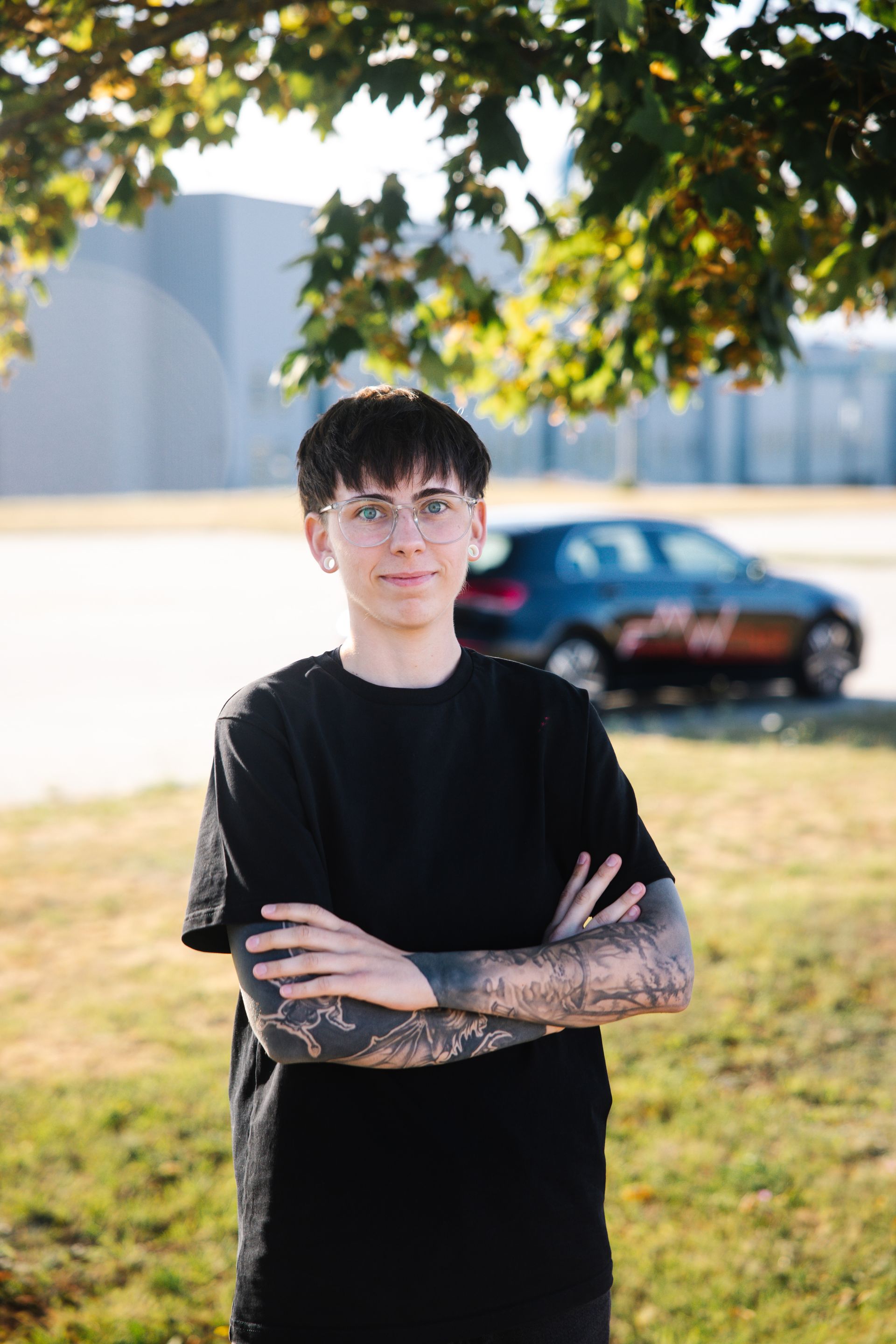 Mann mit Glatze, schwarzem Hemd, verschränkten Armen, lächelt. Auto im Hintergrund. Draußen.