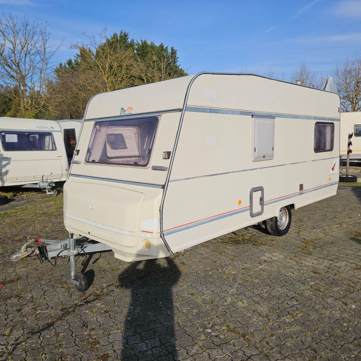 Caravane couleur crème avec attelage, stationnée sur du gravier sur un terrain extérieur.