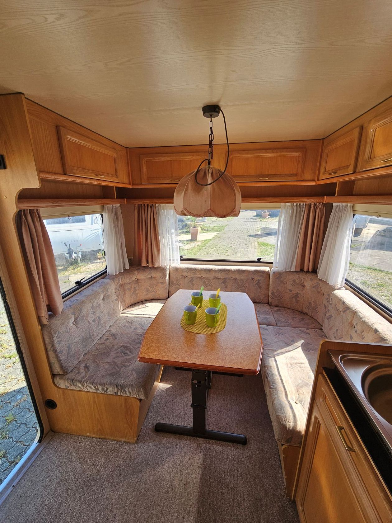Coin repas de camping-car avec lambris, coussins beiges, table en bois, fenêtres et suspension lumineuse.
