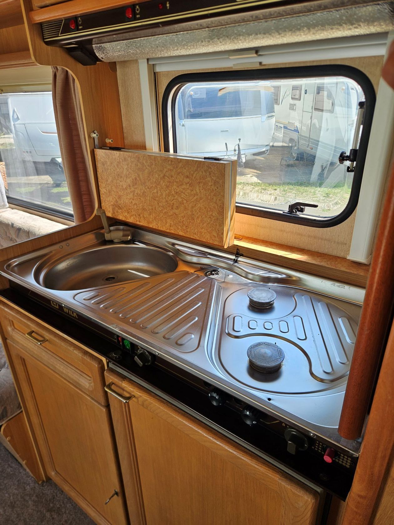 Kitchenette compacte pour camping-car avec évier, plaque de cuisson à deux feux et placards en bois près d'une fenêtre.