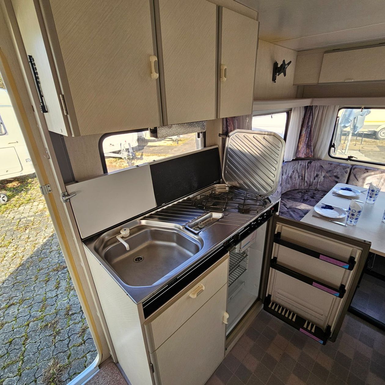 Vue intérieure d'une cuisine de camping-car avec évier, cuisinière, placards et petit réfrigérateur.