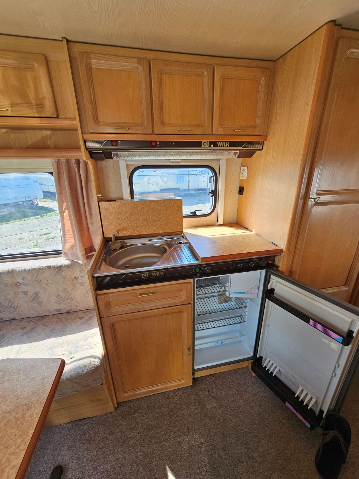 Petite cuisine de camping-car avec placards en bois, évier, plan de travail, cuisinière et fenêtre près de la banquette.