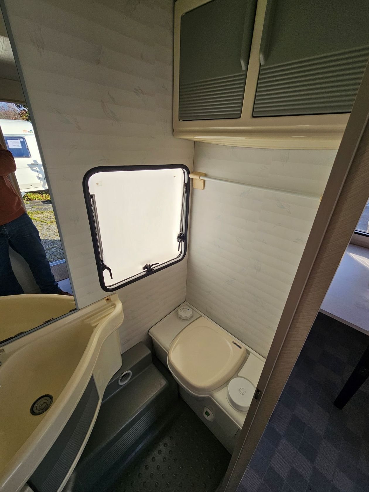 Petite salle de bain de camping-car avec toilettes, lavabo et fenêtre. Tons blancs et gris.