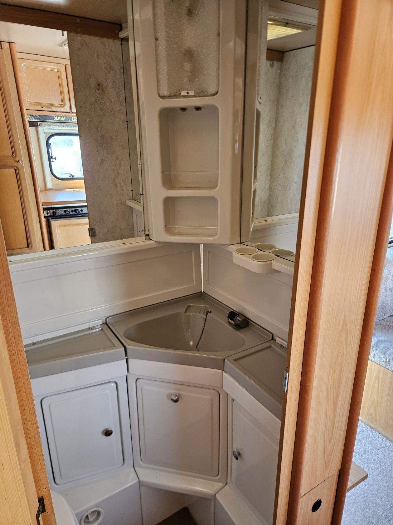 Petite salle de bain blanche pour camping-car avec lavabo d'angle, armoire à miroir et étagères de rangement compactes.