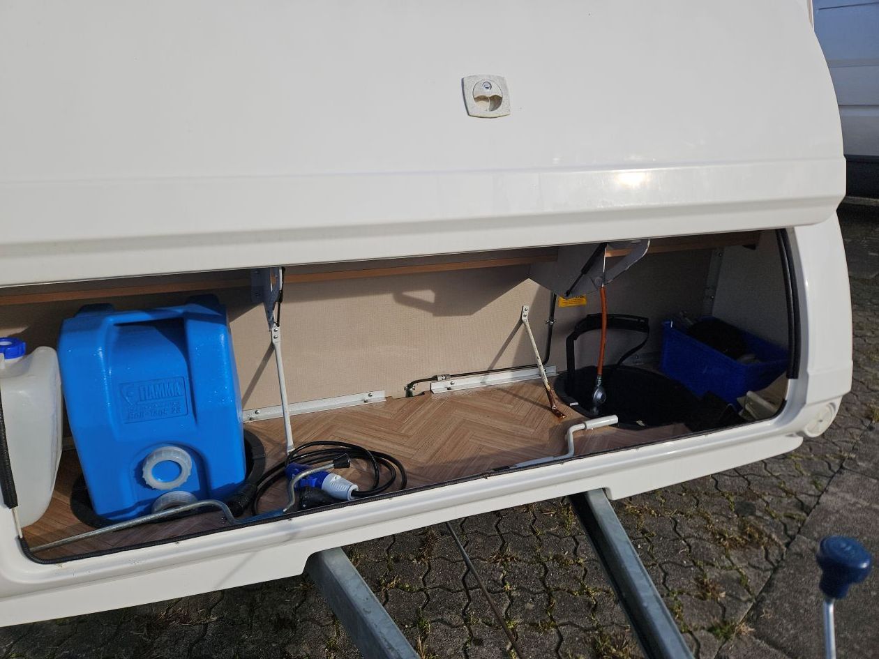 Compartiment de rangement ouvert d'une caravane blanche ; on y voit des bidons d'eau bleus et des câbles d'alimentation.