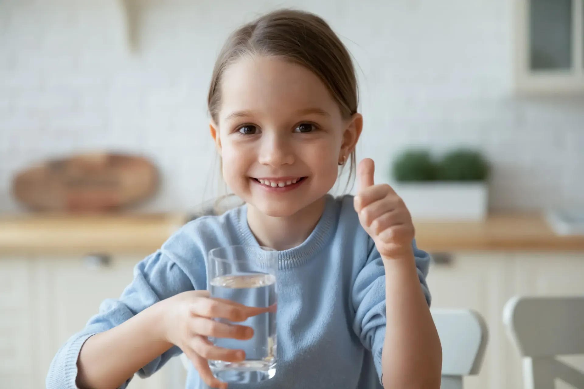 Niña sonriendo, sosteniendo un vaso de agua, dando pulgares hacia arriba en una cocina.