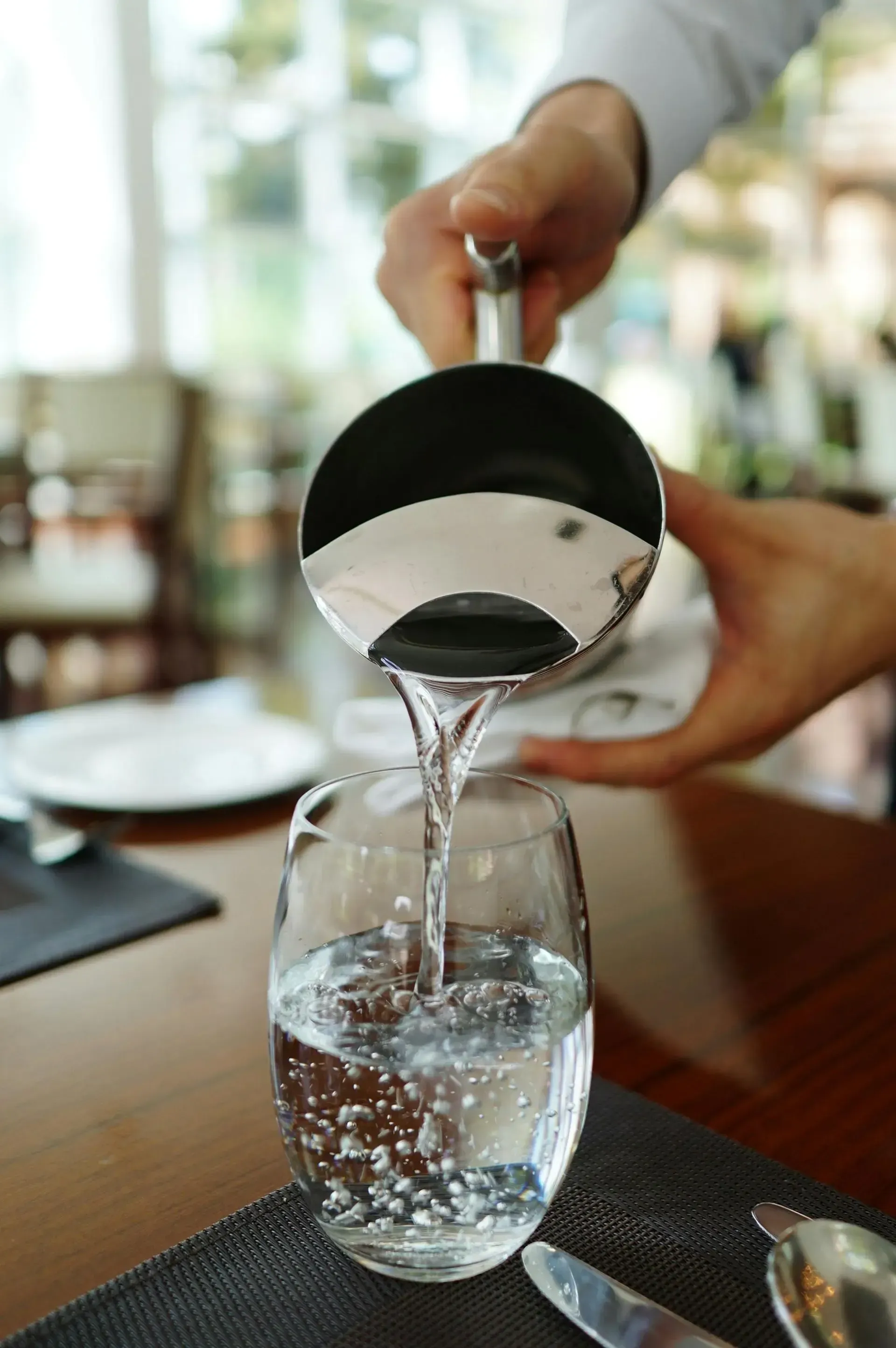 Agua vertida desde una jarra negra a un vaso transparente sobre una mesa de comedor.