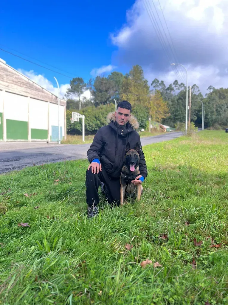 Hombre arrodillado junto a un perro en un campo de hierba, con chaqueta negra. Cielo azul y árboles al fondo.