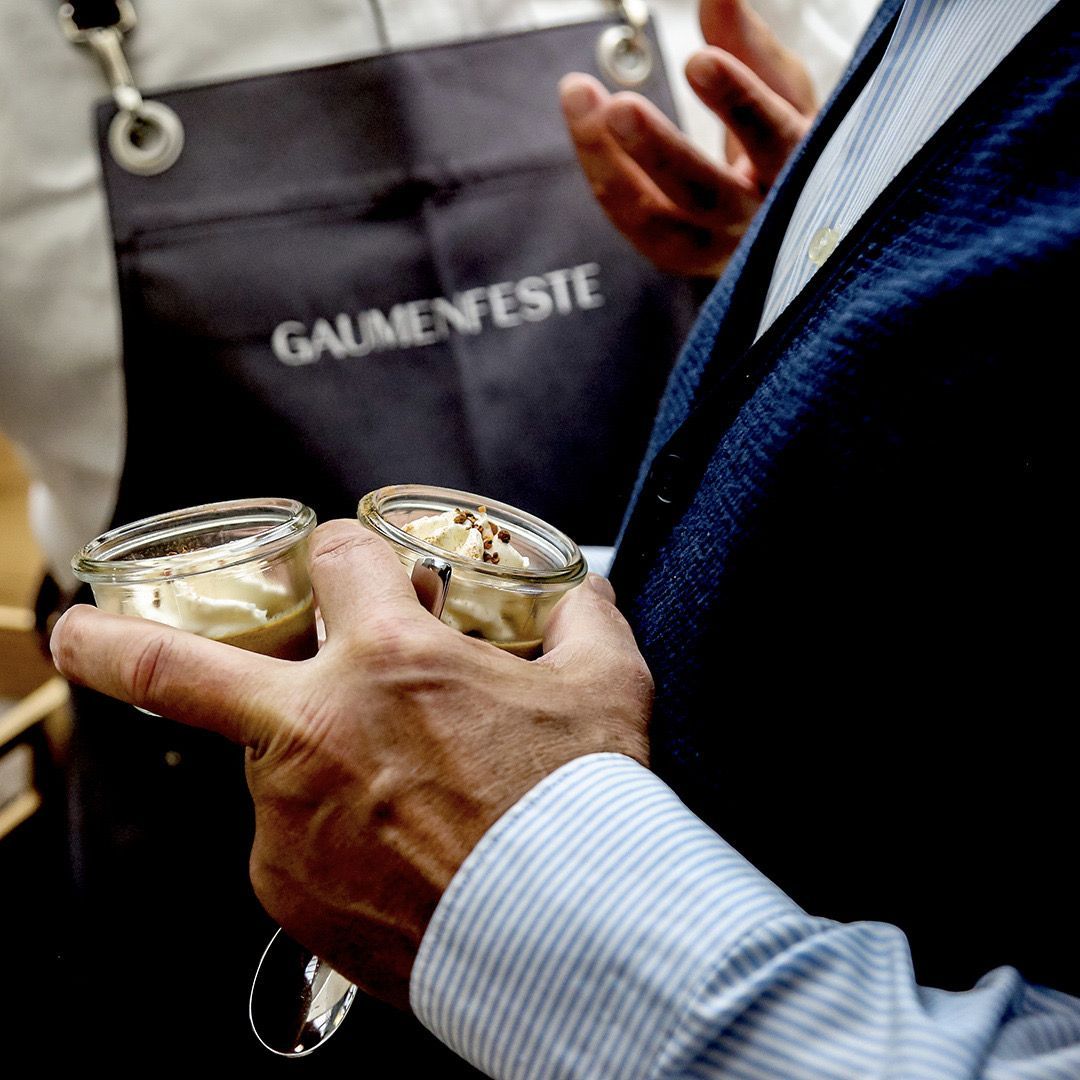 Person hält zwei Desserts im Glas in der Hand, dahinter eine Person mit Schürze