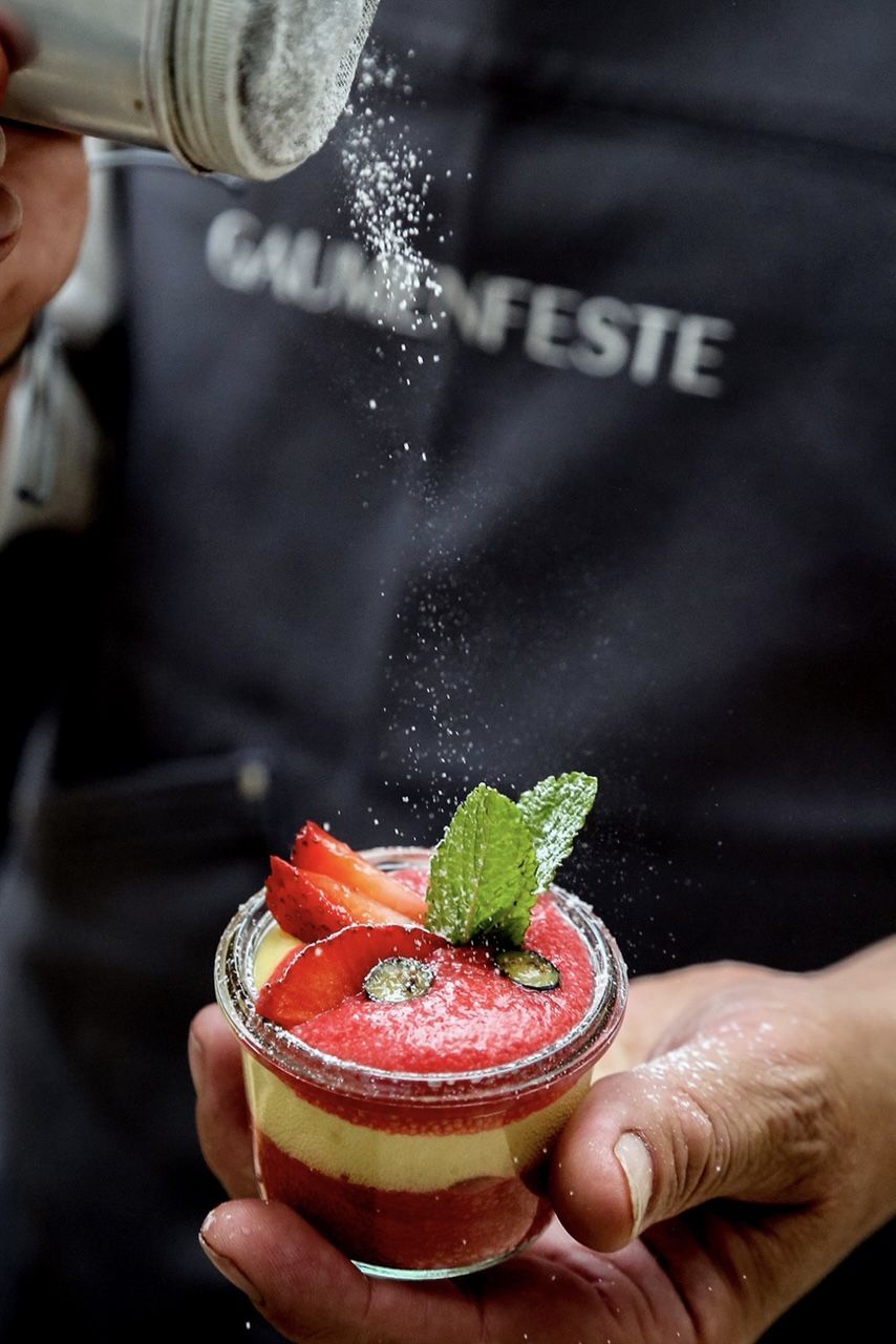 Person streut Puderzucker über ein mit Erdbeeren dekoriertes Schichtdessert im Glas