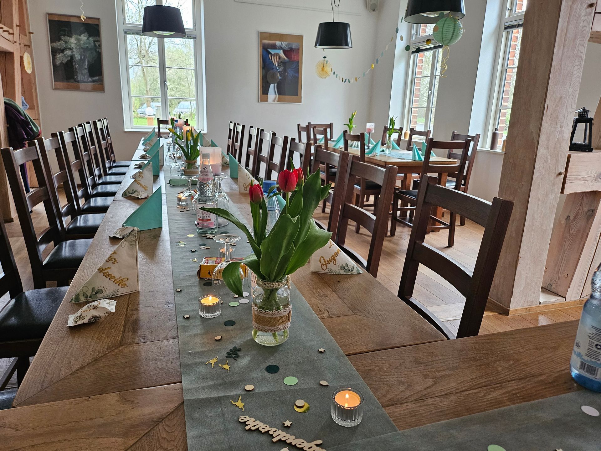 Ein langer, rustikaler Esstisch, gedeckt mit Kerzen und Blumen, steht in einem hellen Raum mit großen Fenstern.