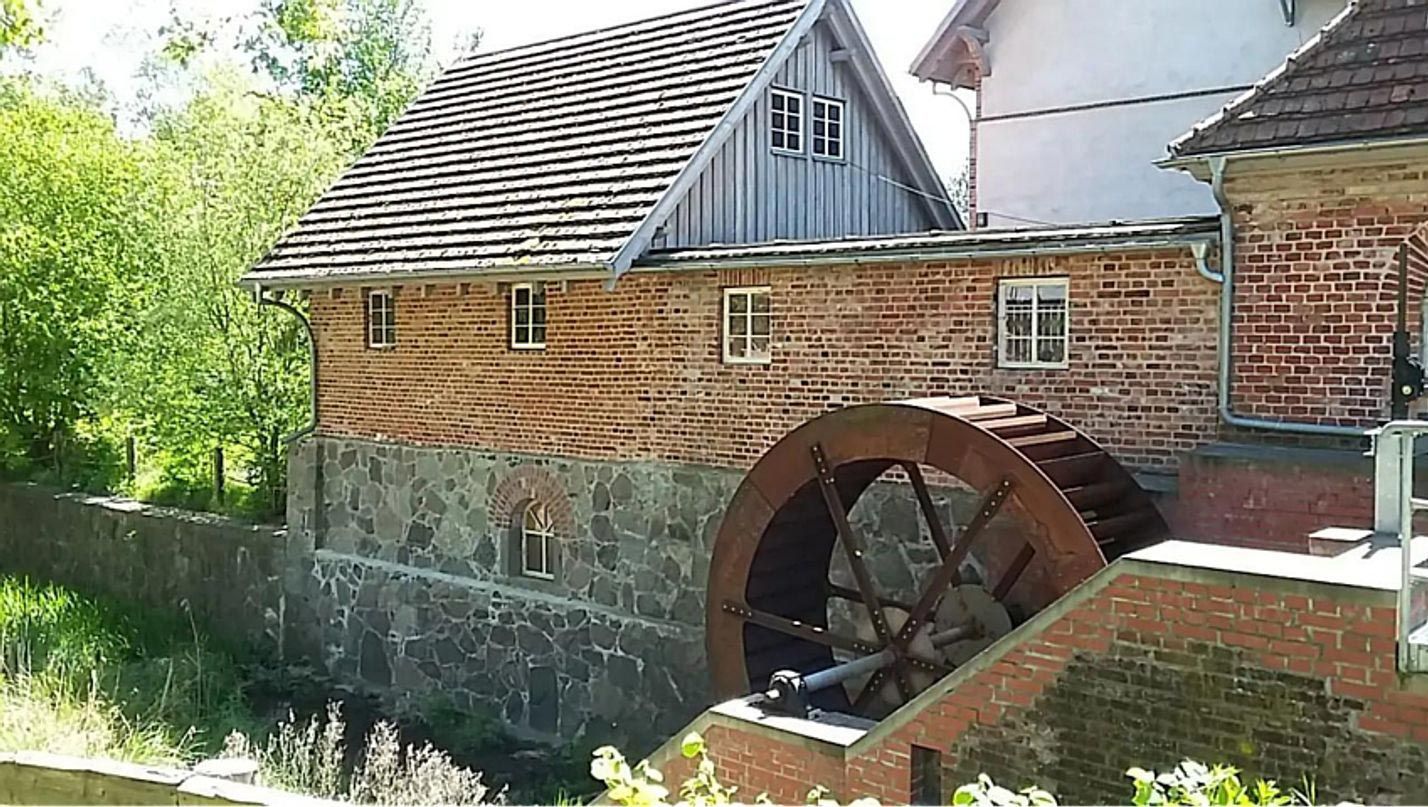Backsteingebäude mit einem großen hölzernen Wasserrad neben einem Steinsockel und einem schrägen Schindeldach