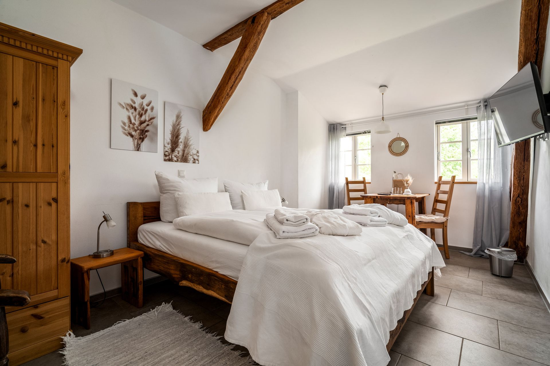 Gemütliches Schlafzimmer mit Holzbett, weißer Bettwäsche, rustikalen Holzbalken und einer Essecke am Fenster