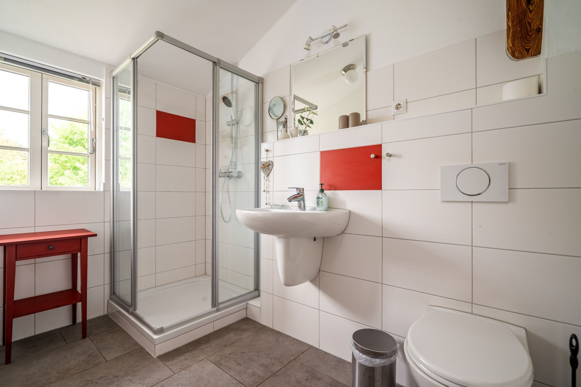 Helles Badezimmer mit Dusche, Waschbecken, Toilette, roten Akzenten und einem Fenster auf der linken Seite.
