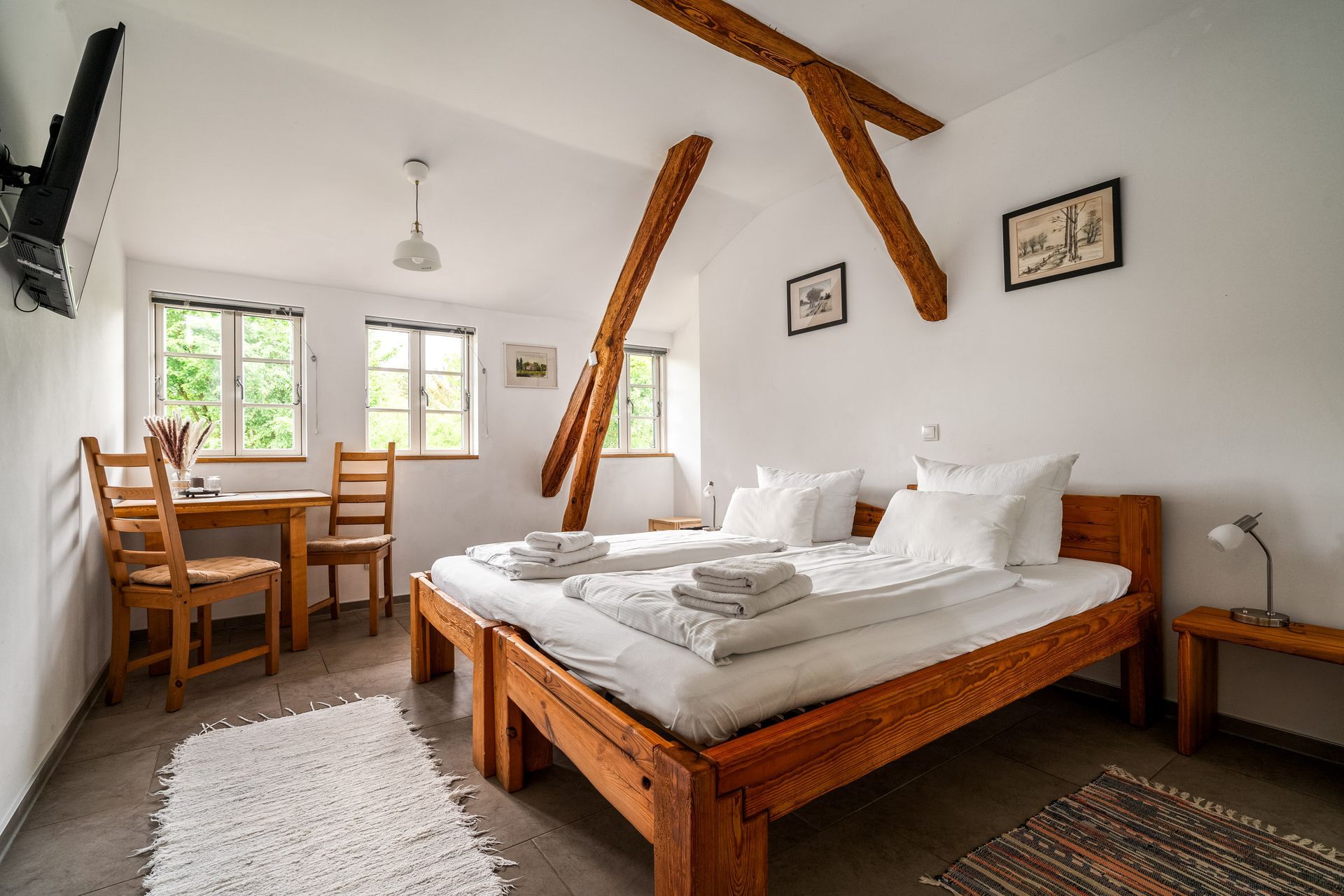 Gemütliches Dachgeschosszimmer mit Holzbalken, einem großen Bett, einem Schreibtisch am Fenster und weißen Wänden